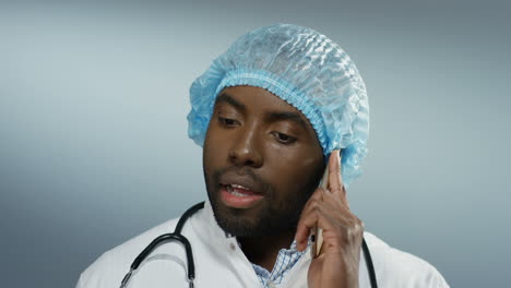 African-American-young-handsome-man-doctor-in-a-hat-and-with-a-stethoscope-on-heck-talking-on-the-mobile-phone-and-solving-some-problem-and-telling-how-to-treat.-Close-up.